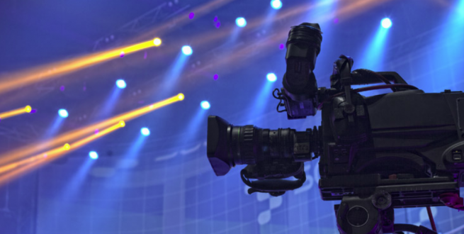 Image of video camera in foreground and a colorfully lit stage in the background.