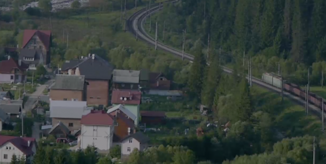 Train moving cargo past a small town near a forest.