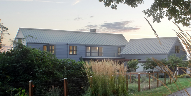 Image of grey home with vegetation and a fence in the foreground.