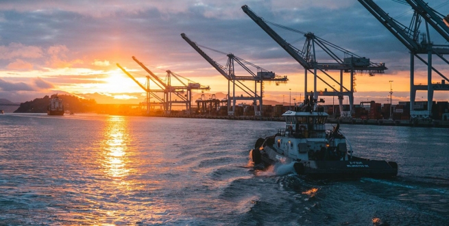 Tugboat driving past cranes in a ship yard at sunset